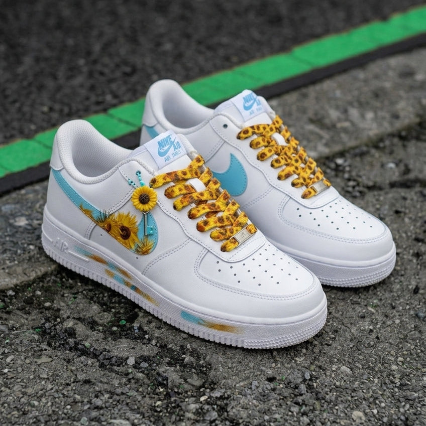 A pair of white Nike Air Force 1 Low sneakers with custom yellow and blue designs, including sunflower graphics on the side and leopard-print laces, resting on a gray asphalt surface.